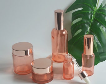 Puede incluir: Colección de envases cosméticos de vidrio color melocotón con tapas de oro rosa. El conjunto incluye frascos, pulverizadores y un frasco cuentagotas. Los envases están dispuestos sobre una superficie blanca con una gran hoja verde de fondo.