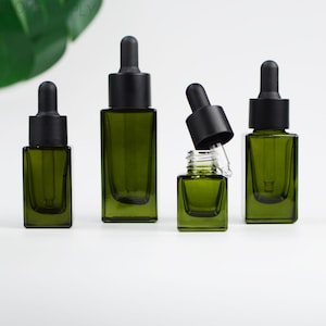 Könnte beinhalten: Vier grüne Glas-Tropfflaschen mit schwarzen Kappen sind auf einer weißen Oberfläche angeordnet. Die Flaschen sind unterschiedlich groß, eine ist geöffnet und zeigt eine Glastropfpipette. Die rechteckigen Flaschen haben eine dunkelgrüne Tönung.