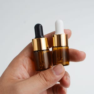 May include: Two small amber glass dropper bottles with gold-colored collars. One bottle has a black dropper, the other a white dropper. The bottles are held in a hand against a white background. The bottles are likely for essential oils or cosmetic products.