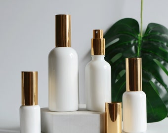 May include: A collection of white glass bottles with gold-colored caps and spray tops. The bottles vary in size and shape, suitable for essential oils or perfumes. The image is set against a neutral background with a large green leaf.