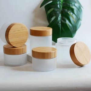 Puede incluir: Seis tarros de vidrio esmerilado con tapas de bambú. Los tarros son de varios tamaños y están dispuestos sobre una superficie blanca.