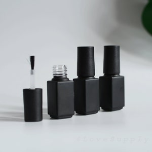Op de afbeelding: Een verzameling zwarte matte nagellakflesjes met bijpassende doppen. Eén flesje is open en toont een helder glazen interieur en een kwastapplicator. De andere flesjes zijn gesloten. De flesjes staan op een witte ondergrond.