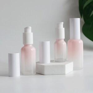 May include: Four glass bottles with white pump tops and a gradient pink to clear design. The bottles are on a white hexagonal platform.