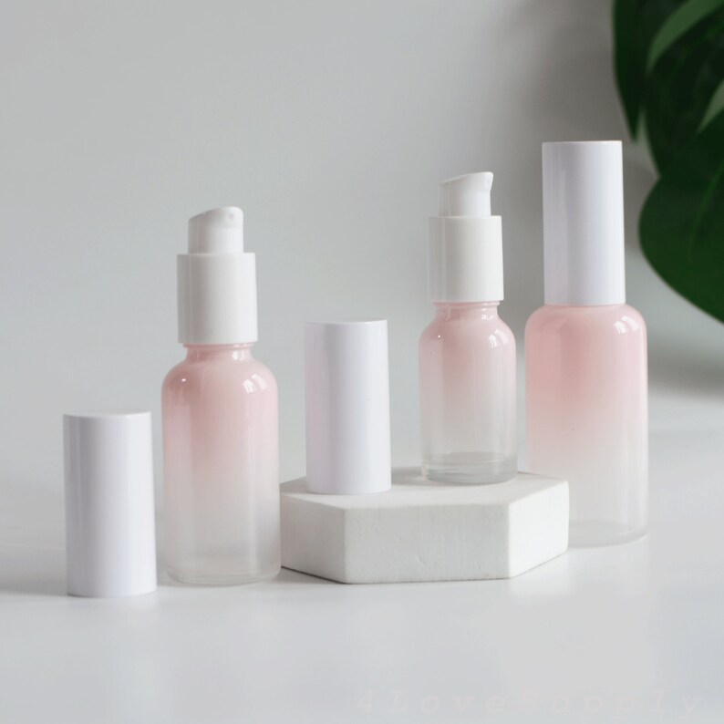 May include: Four glass bottles with white pump tops and a gradient pink to clear design. The bottles are on a white hexagonal platform.