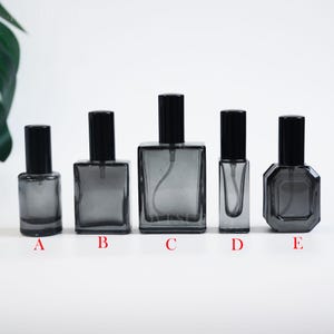 May include: Five empty glass spray bottles with black caps, labelled A through E. Bottle A is cylindrical, B is rectangular, C is a large rectangular bottle, D is a slim rectangular bottle, and E is an octagonal bottle. All are dark grey.