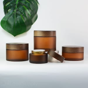 Könnte beinhalten: Eine Sammlung von bernsteinfarbenen Glasbehältern mit Holzdeckeln. Die Gläser sind unterschiedlich groß und auf einer weißen Oberfläche angeordnet. Ein Glas hat seinen Deckel abgenommen, wodurch ein weißes Innenleben sichtbar wird. Ein großes grünes Blatt ist im Hintergrund zu sehen.