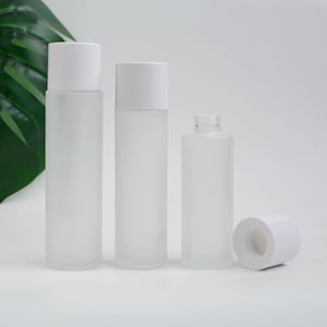 May include: Three frosted glass bottles with white caps and a detached cap. The bottles are cylindrical and vary in height. The background is white, with a green leaf visible on the left side.