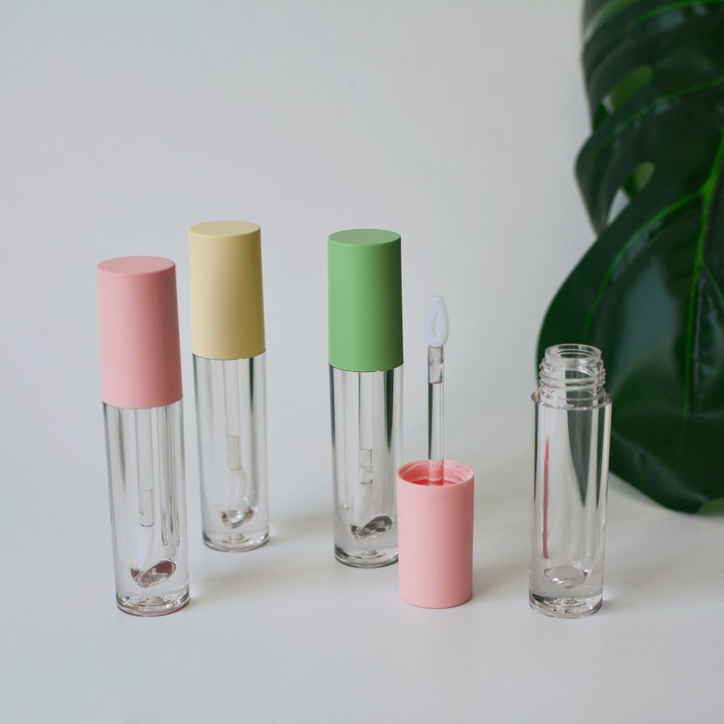 May include: Four clear lip gloss tubes with different colored caps: pink, yellow, green, and pink. The tubes are on a white surface with a green leaf in the background.