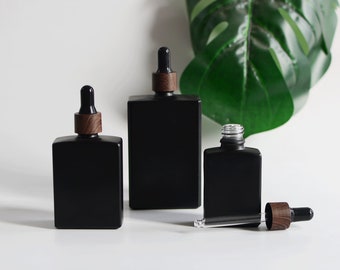 May include: Three black square glass bottles with wooden-look caps and droppers. One bottle is open, with the dropper lying next to it. The bottles are of varying sizes. The background is white with a large green leaf.