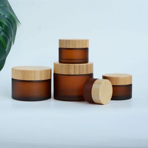 May include: A collection of amber-colored glass jars with bamboo lids. The jars are in various sizes, arranged together. The jars have a frosted appearance. The lids are made of light-colored bamboo. The jars are suitable for storing cosmetics or other small items.