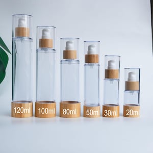 Puede incluir: Botellas de bomba cilíndricas transparentes con bases y tapas de madera. Los tamaños varían de 20 ml a 120 ml, con etiquetas que indican el volumen. Las botellas tienen un cuerpo de plástico transparente y un dispensador de bomba blanco.