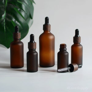 May include: Five amber glass bottles with black dropper tops and wooden caps. The bottles are frosted and have a matte finish. The bottles are arranged in a row on a white surface.