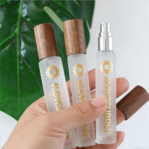 Könnte beinhalten: Drei Sprühflaschen aus mattiertem Glas mit Holzdeckeln. Jede Flasche hat ein goldenes Sonnenblumenmuster und den Text "4LOVESUPPLY". Die Flaschen werden in einer Hand vor einem Hintergrund aus grünen Blättern gehalten.
