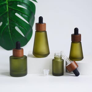 Puede incluir: Cuatro frascos cuentagotas de vidrio verde oliva mate con collares de madera y bulbos de goma negros. Dos frascos son altos y cónicos, uno es corto y cilíndrico, y uno está abierto con una pipeta de vidrio y tapa de madera separadas.
