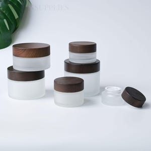 Puede incluir: Una colección de tarros cosméticos de vidrio esmerilado con tapas de madera marrón oscuro. El conjunto incluye varios tamaños, con algunos tarros apilados y uno mostrado sin tapa, revelando un revestimiento interior de plástico blanco.