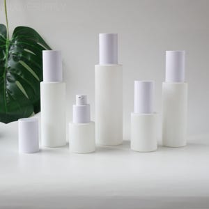 May include: A selection of white frosted glass bottles with white caps and a pump dispenser. The bottles are of varying sizes and shapes, suitable for cosmetic products. The background is a plain white surface with a green leaf.