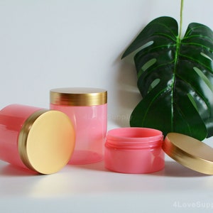 Pode incluir: Três frascos de plástico rosa com tampas douradas. Os frascos estão vazios e têm um acabamento liso e brilhante. As tampas são brilhantes e têm uma superfície ligeiramente texturizada.