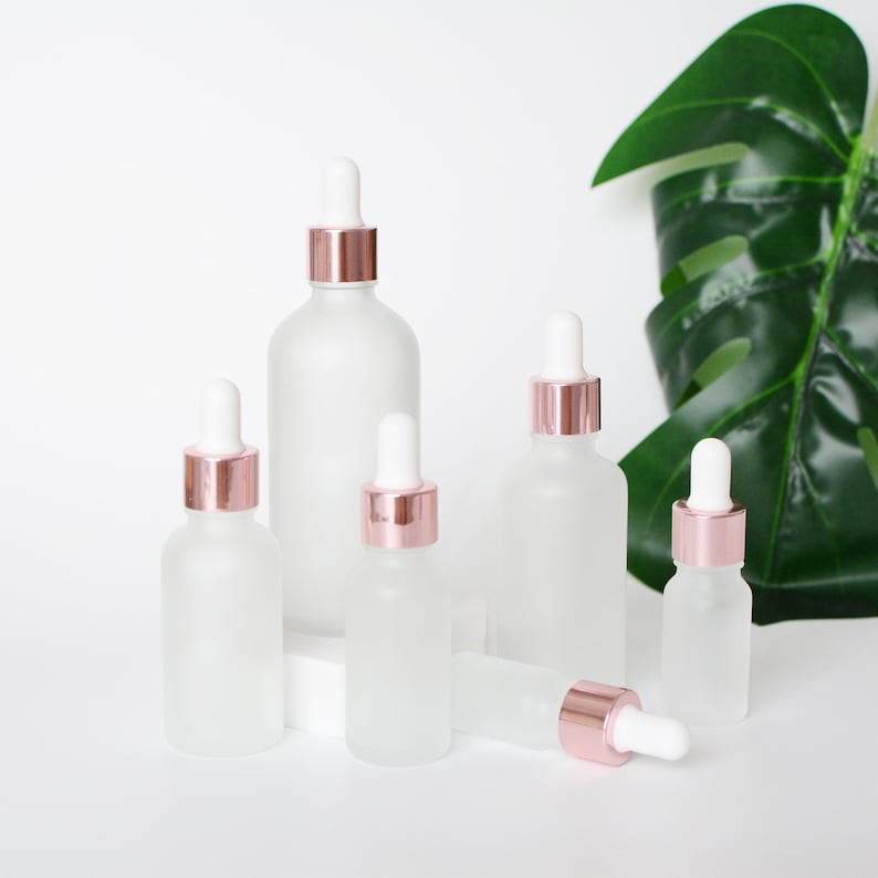 May include: A collection of frosted glass dropper bottles with rose gold collars and white droppers. The bottles vary in size and are arranged on a white surface, with a large green leaf in the background.