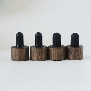 Puede incluir: Cuatro tapones de botella de madera con tapas de goma negra. Los tapones tienen un acabado de vetas de madera oscura y una forma cilíndrica. Las tapas de goma negra son redondeadas y lisas.