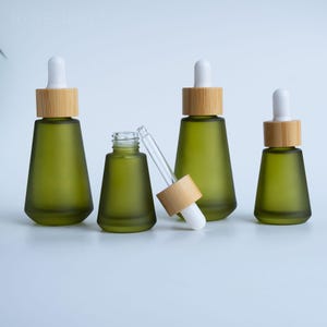 Puede incluir: Cuatro frascos cuentagotas de vidrio verde con tapas de bambú y cuentagotas blancos. Un frasco está abierto, con el cuentagotas parcialmente insertado. Los frascos están dispuestos sobre una superficie blanca.