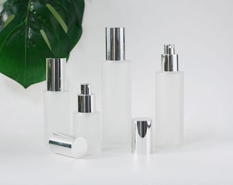 May include: Four frosted glass bottles with silver caps and pumps. The bottles are empty and are arranged in a row on a white surface.