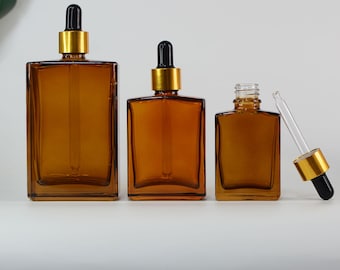 May include: Three amber glass dropper bottles of varying sizes. Each bottle has a rectangular shape with a gold collar and a black dropper. The bottles are arranged from largest to smallest.