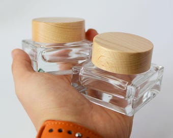 May include: Two clear glass square jars with wooden lids. The jars are empty and are being held in a hand.