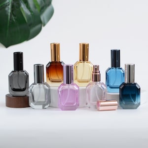 May include: A collection of perfume bottles in various colors, including clear, gray, amber, yellow, pink, blue, and dark blue. Each bottle has a geometric shape and a metallic cap. Some bottles have spray nozzles, and one has a detached rose gold cap.