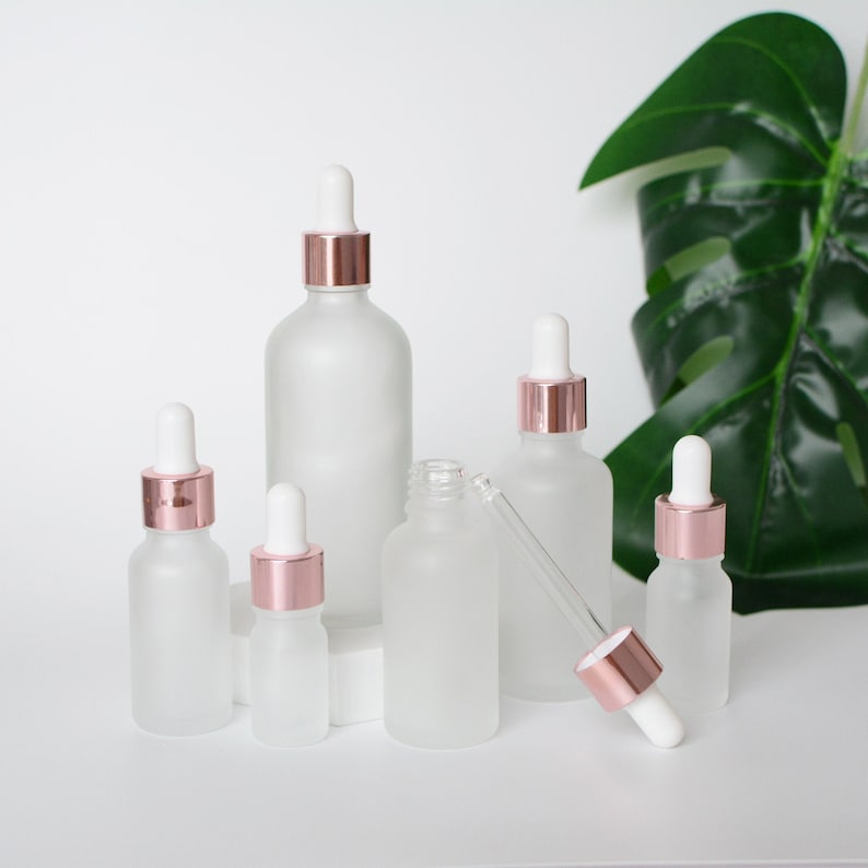 May include: A collection of frosted glass dropper bottles with rose gold collars and white droppers. The bottles vary in size, with one bottle open and a dropper separate. A green leaf is in the background.