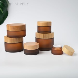 May include: A collection of amber-colored glass jars with bamboo lids. The jars vary in size, with a frosted finish. The lids are made of light-colored bamboo. The jars are arranged on a white surface. The jars are suitable for storing cosmetics or other small items.