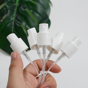 Op de afbeelding: Een verzameling witte plastic sproeipomptoppen met doorzichtige buisjes. De sproeiers worden in een hand gehouden, met een groene, bladerrijke achtergrond. Deze zijn waarschijnlijk voor cosmetica- of reinigingsmiddelenflessen.