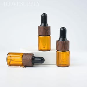 May include: Amber glass dropper bottles with dark wood grain collars and black droppers. One bottle is lying on its side, while two are upright. The bottles are on a white surface.