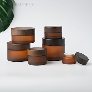 May include: A selection of amber glass jars with wooden lids. The jars are of varying sizes and are grouped together. The lids are a dark brown colour. The jars are suitable for storing cosmetics or other small items.