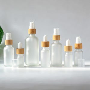 May include: A selection of frosted glass bottles with bamboo lids and spray or dropper applicators. The bottles are in various sizes and shapes, ideal for essential oils or cosmetics. The bottles are arranged on a white surface.