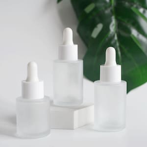 Könnte beinhalten: Drei milchglasflaschen mit weißen Tropferaufsätzen. Die Flaschen sind leer und stehen auf einer weißen Oberfläche.