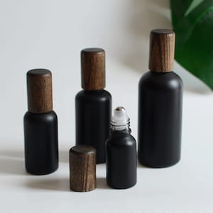 May include: A selection of black glass bottles with wooden caps. The bottles are of varying sizes, including a roller bottle with a silver rollerball. The wooden caps have a natural wood grain finish. The bottles are displayed on a white surface.