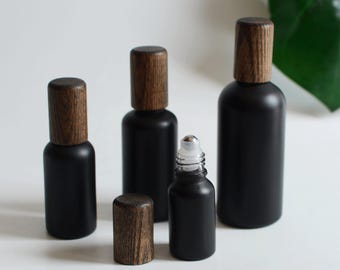 May include: A collection of black glass bottles with wooden caps. The bottles vary in size, including a roller bottle with a silver rollerball. The wooden caps have a natural wood grain finish. The bottles are arranged on a white surface.