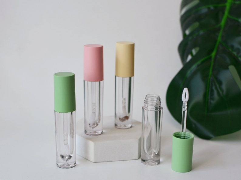May include: Four clear lip gloss tubes with different colored caps. The caps are pink, yellow, green, and green. The tubes are on a white surface.