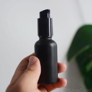 Puede incluir: Un frasco cilíndrico de color negro mate que cuenta con un mecanismo de bomba dosificadora de plástico negro en la parte superior. El frasco tiene un acabado liso y opaco con un diseño de hombros redondeados, ofreciendo un recipiente funcional.