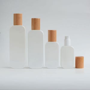 May include: A selection of white, rectangular bottles with rounded corners and wooden caps. The bottles are of varying sizes, with one featuring a pump dispenser. The wooden caps are a light brown colour. The bottles are arranged on a white surface.