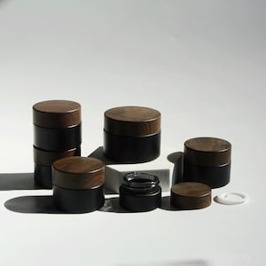 May include: A set of five black glass jars with wooden lids. The jars are of different sizes and are arranged on a white surface.