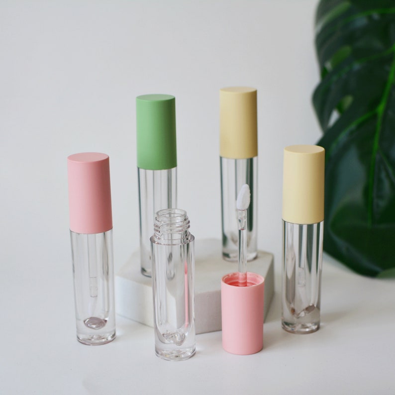 May include: Five clear lip gloss tubes with different colored caps. The caps are pink, green, yellow, and a lighter shade of yellow. The tubes are on a white surface.