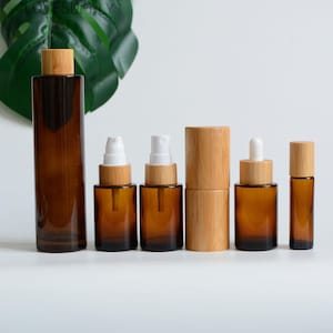 May include: An assortment of amber glass bottles and jars, each with a bamboo lid. The set includes a tall rectangular bottle, spray bottles, a dropper bottle, and a roll-on bottle. The containers are arranged on a white surface, suggesting a skincare or essential oil product line.