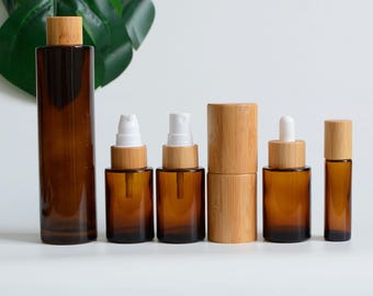 May include: A collection of amber glass bottles and jars with bamboo lids. The set includes a tall rectangular bottle, spray bottles, a dropper bottle, and a roll-on bottle. The containers are arranged on a white surface, suggesting a skincare or essential oil product line.