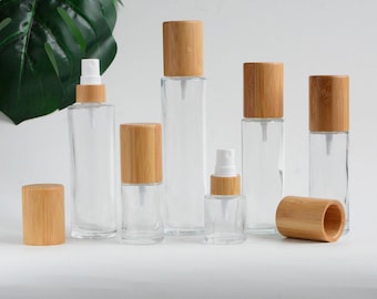 May include: Clear glass spray bottles with natural bamboo lids. The bottles vary in size and shape, with some being tall and rectangular and others shorter and round. The bamboo lids are a light brown color. The bottles are arranged on a white surface.