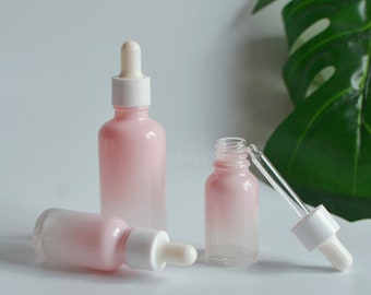 May include: Pink and clear glass dropper bottles with white droppers. The bottles are in various sizes, with a gradient pink color that fades to clear. The bottles are for essential oils or cosmetic products.