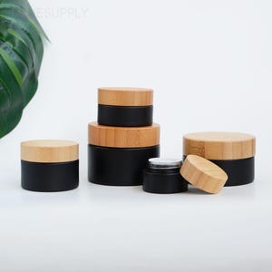 Może przedstawiać: Kolekcja czarnych słoiczków kosmetycznych z naturalnymi bambusowymi wieczkami. Słoiczki różnej wielkości, niektóre ułożone w stos. Wieczka są jasnobrązowe, a słoiczki cylindryczne. Słoiczki są puste.