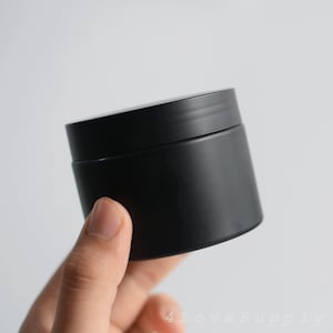 Pode incluir: Um recipiente redondo de plástico preto com uma tampa. O recipiente está sendo segurado em uma mão.