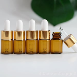 May include: Five amber glass bottles with gold lids and white droppers. The bottles are arranged in a row on a white surface. One bottle is lying on its side with the dropper facing up.