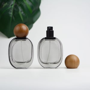 Pode incluir: Dois frascos de perfume ovais transparentes com borrifadores pretos e tampas de madeira. Um frasco tem uma tampa de madeira, e o outro tem um borrifador preto. Uma tampa de madeira solta está ao lado dos frascos.
